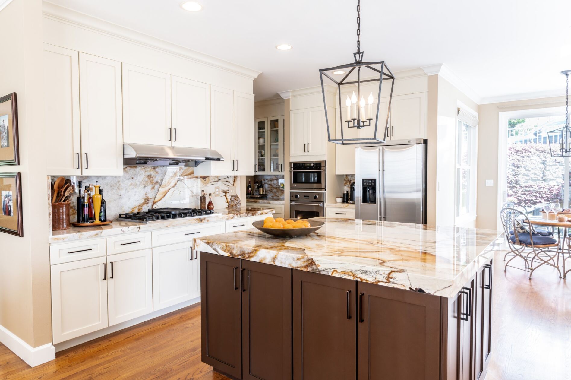 Modern kitchen with white cabinets, stainless steel appliances, marble countertops, a center island with a fruit bowl, and a pendant light fixture. Background shows a dining area with large windows.